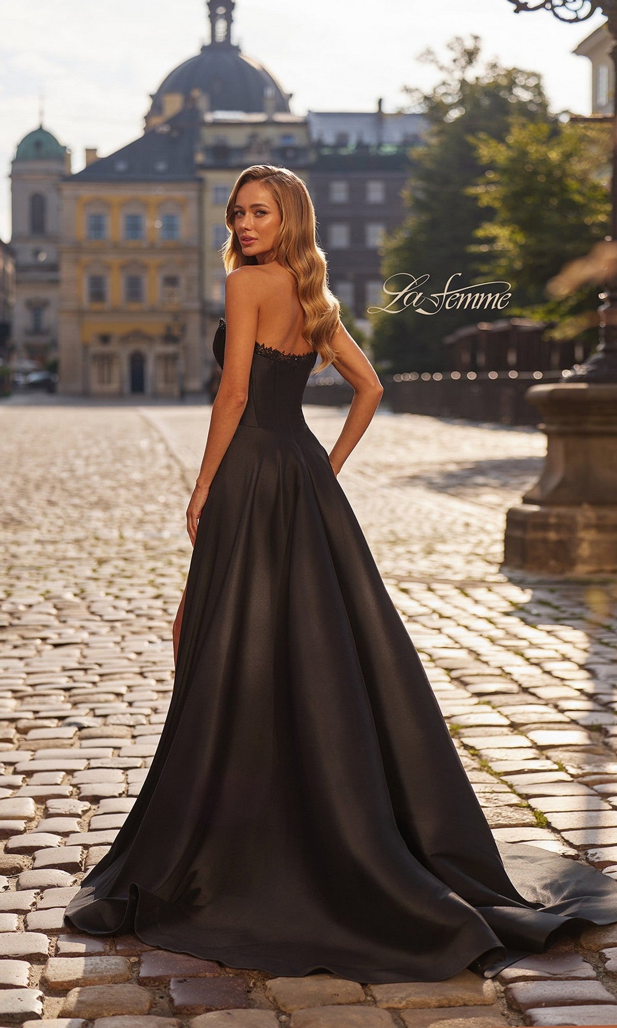 A woman wearing the La Femme 33353 long black Mikado strapless prom dress stands on a cobblestone street, glancing over her shoulder, with historic buildings and a clear sky in the background.